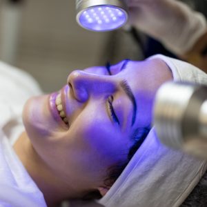 Female Beautician Doing Blue Light Therapy On face of pretty young woman